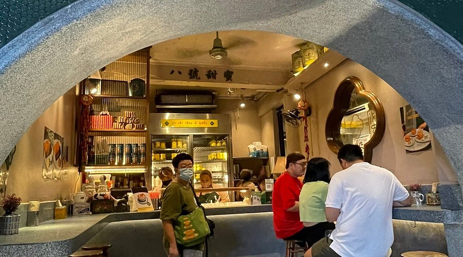 A trendy open-air Chinese dessert shop with a large circular entrance, red neon signs, and customers sitting on wooden stools eating pudding.